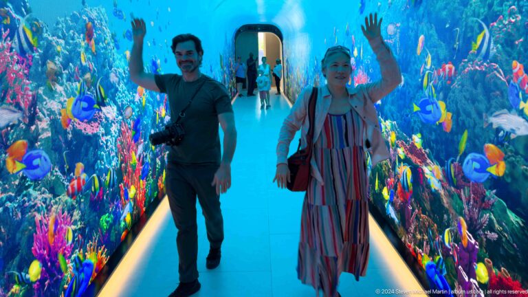 Smiling man with a camera and woman in a striped dress waving in a blue fish corridor.