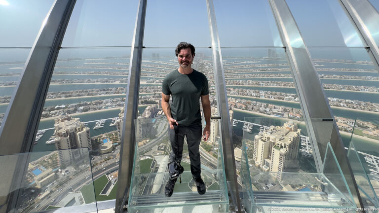 Full-body portrait of a man standing on a glass observation platform above Palm Jumeirah.
