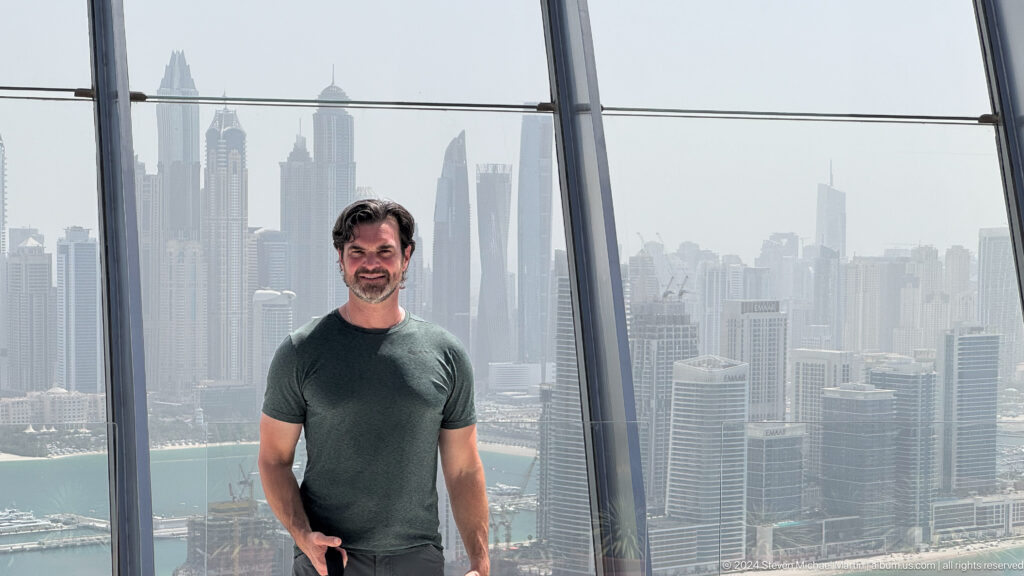 Centered man in a dark green shirt with Dubai Marina towers behind him.