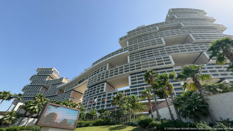 Wide low-angle view of Atlantis The Royal above palms, gardens, flags, and an outdoor screen.