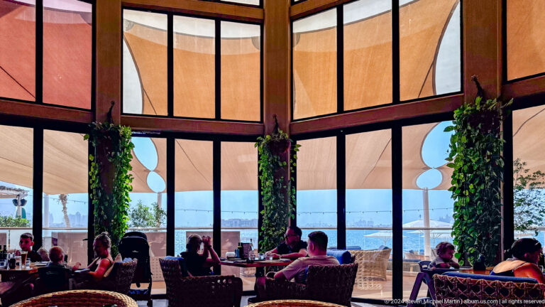 Waterfront restaurant with diners, hanging plants, tall windows, and fabric sunshades.