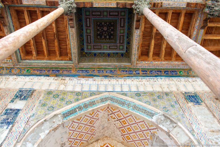 Two wooden columns framing painted ceiling panels in a mosque portico.