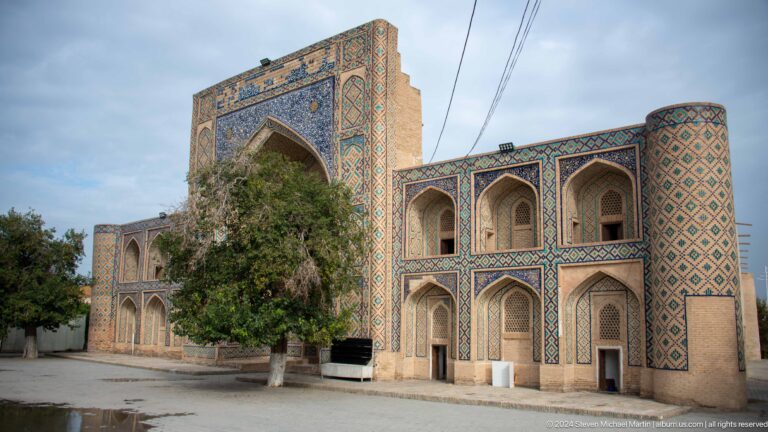 Tiled two-story madrasa façade partly screened by a large tree.