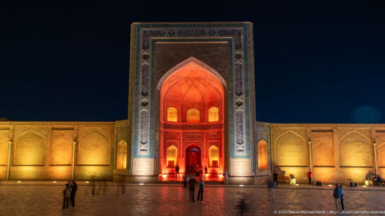 Red-lit monumental portal reflected on paving with visitors below.