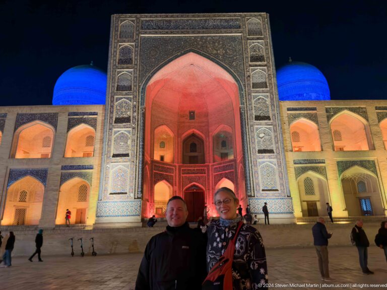 Two people posing before a red-lit madrasa portal and blue domes at night.