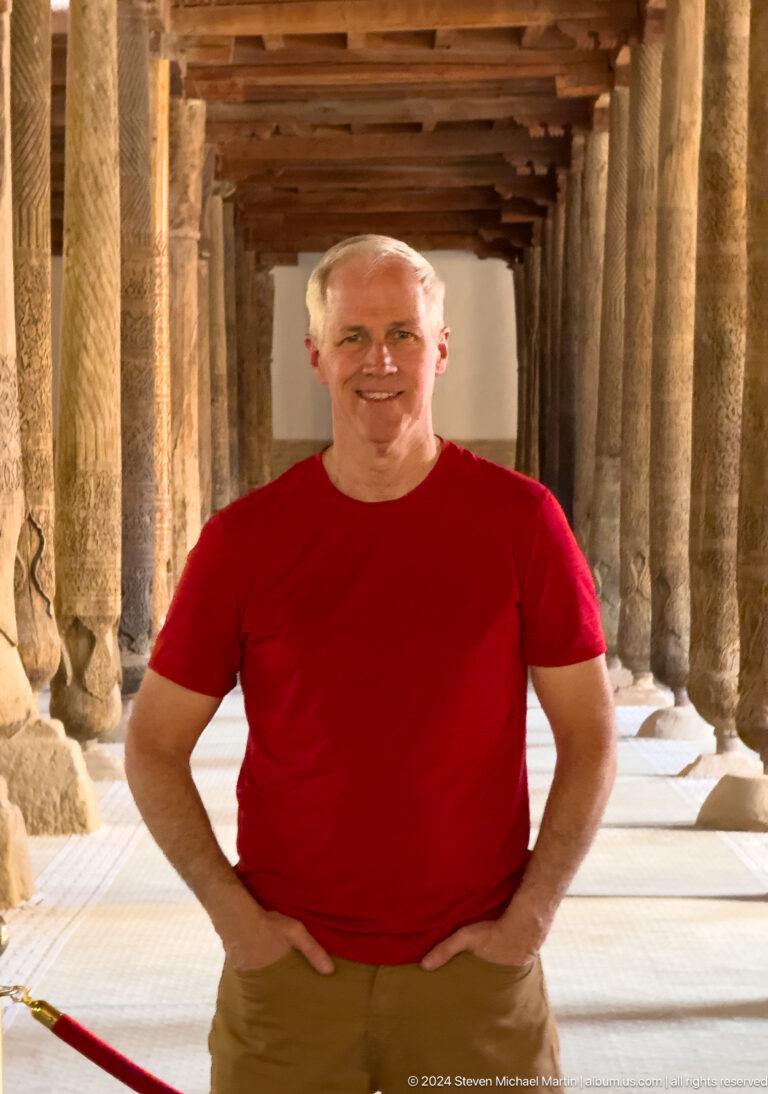 Smiling man in red shirt among juma mosque columns.