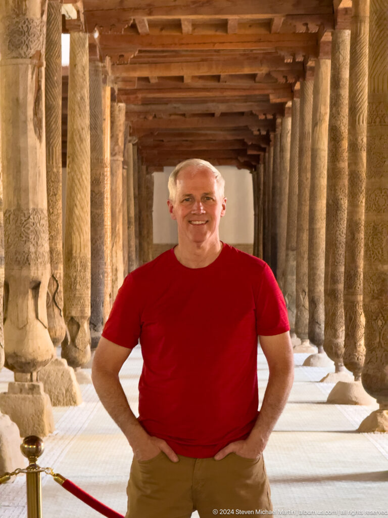 Smiling man in red shirt among wooden mosque columns.