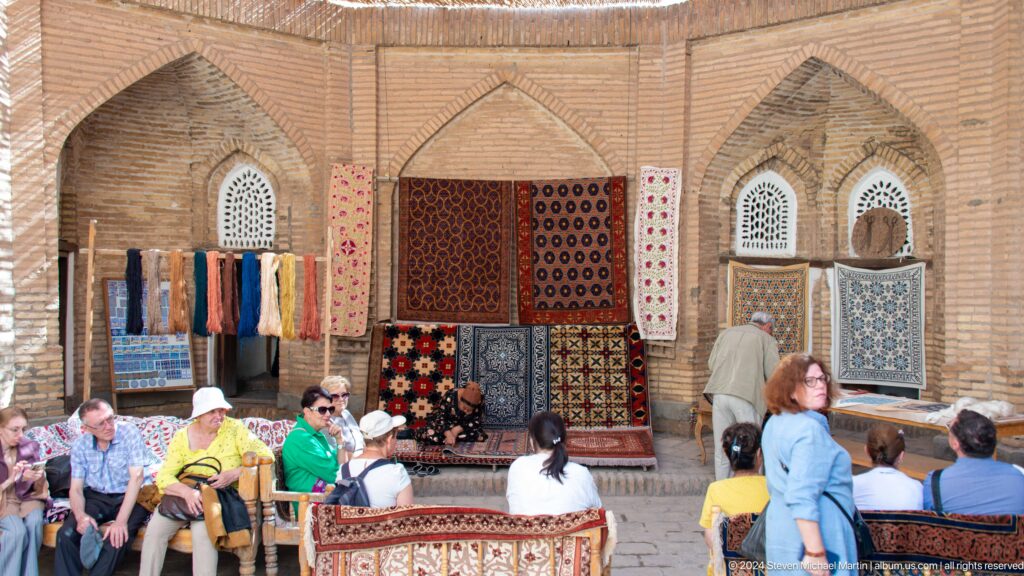 Visitors examining carpets and textiles displayed on a madrasa wall.