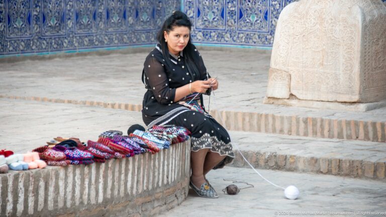Woman seated beside embroidered goods in palace courtyard.