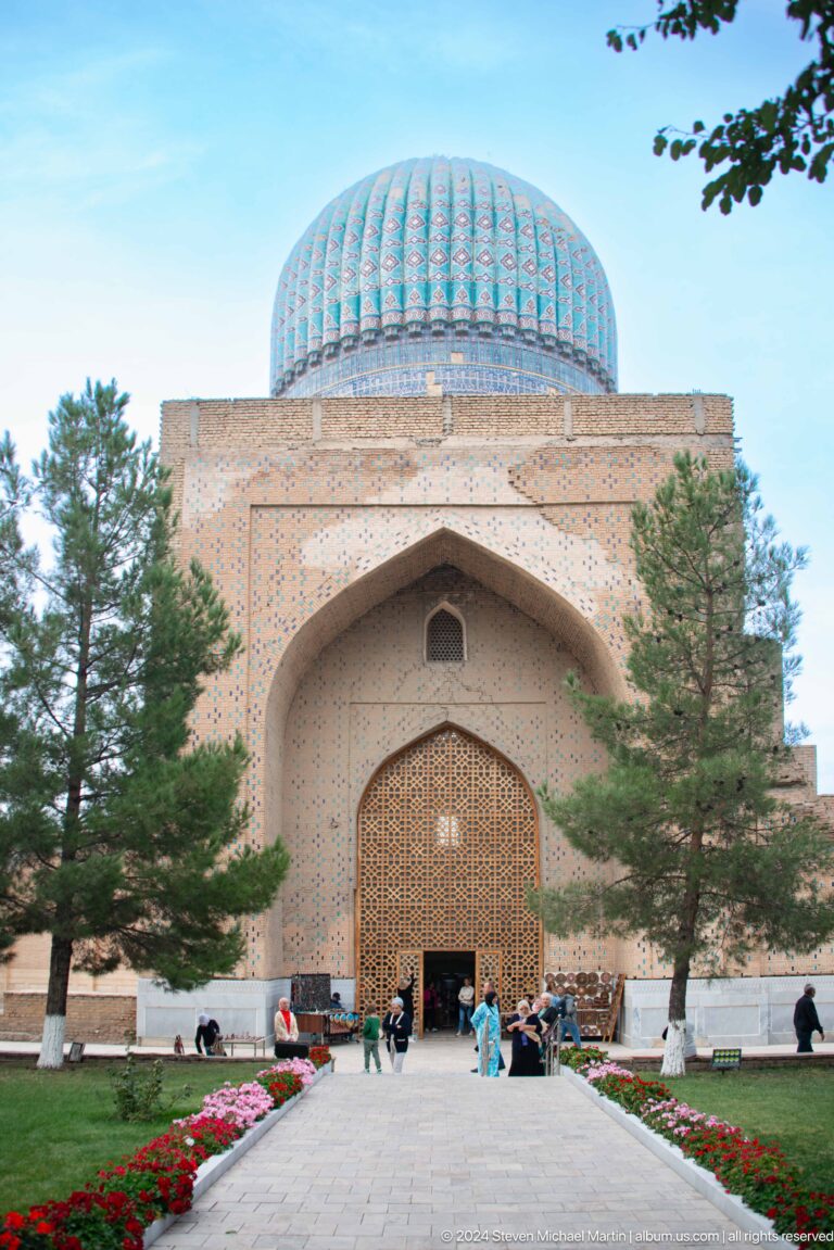 Brick mosque façade with a pointed recessed portal, wooden lattice screen, trees, and turquoise dome above.