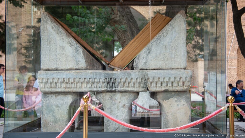 Large pale stone Quran stand with sloping supports, displayed behind glass and a red rope barrier.