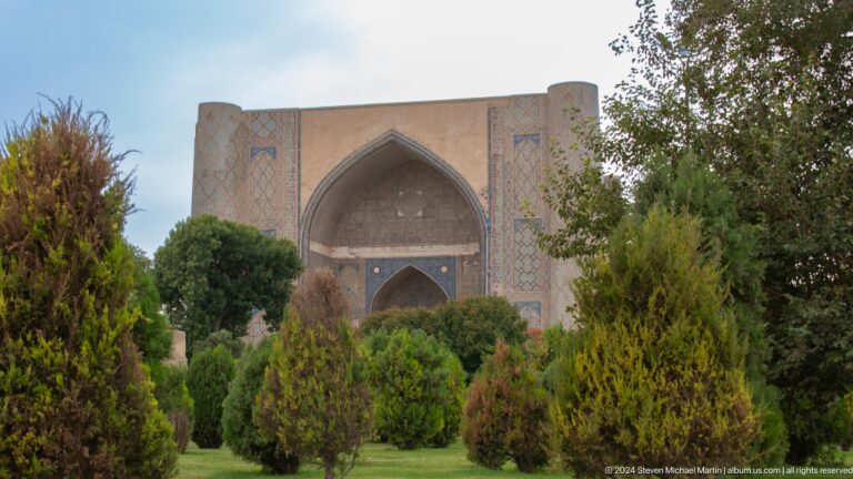 Distant monumental mosque iwan framed by dense green trees in a landscaped courtyard.