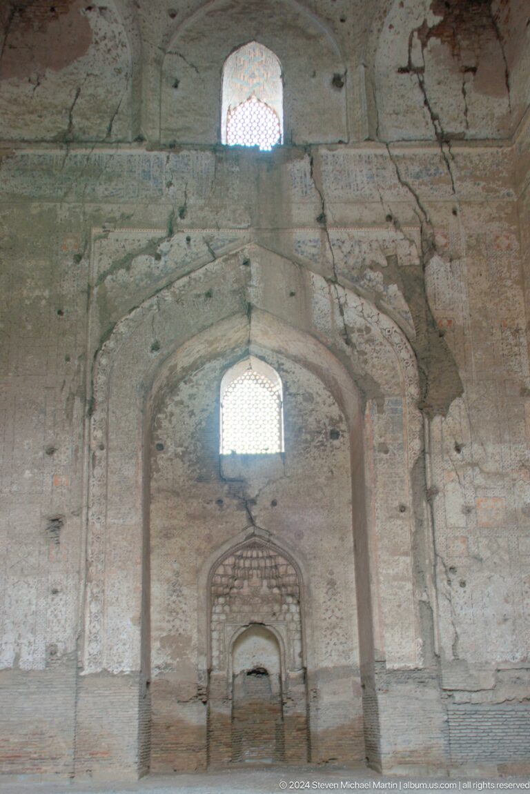 Dim ruined interior with a pointed vault, bright upper window, central niche, and worn masonry surfaces.