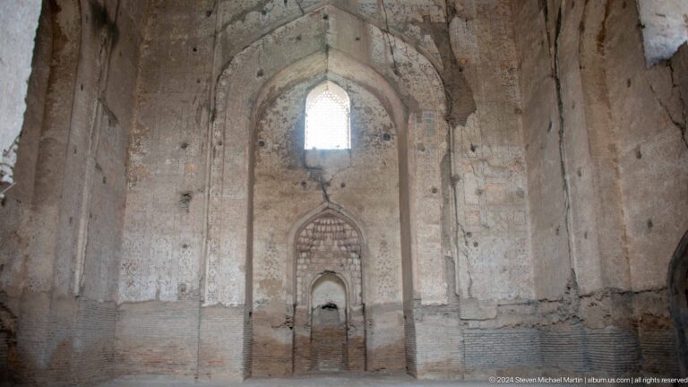 Wide symmetrical ruined interior with a high pointed window, stacked niches, exposed brick, and worn plaster.