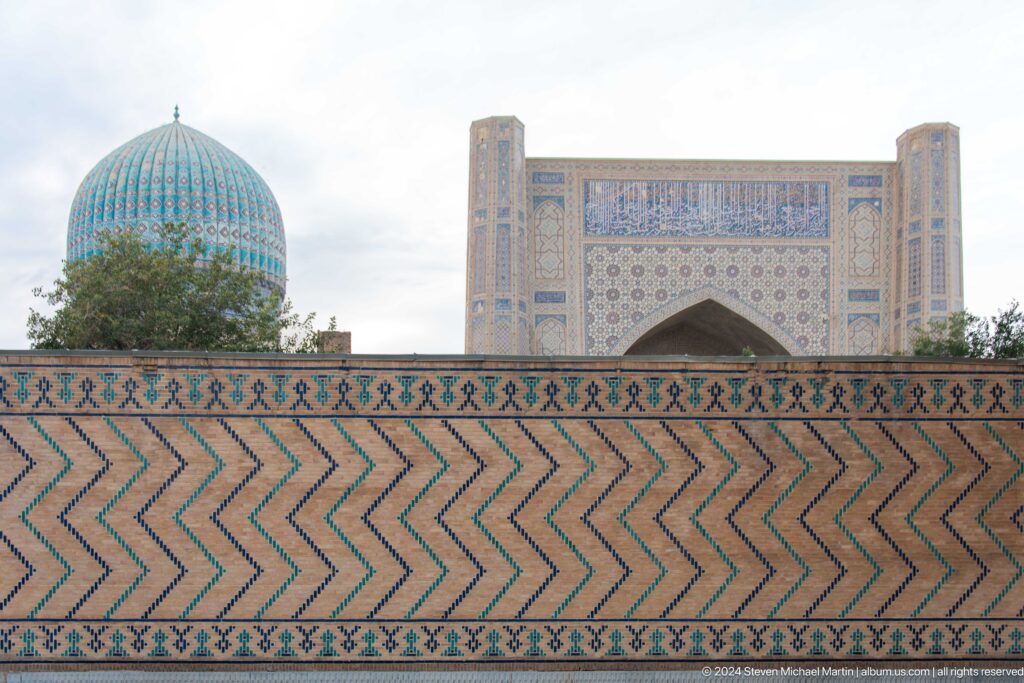 Chevron-patterned tiled wall in front of a turquoise dome and monumental mosque iwan.