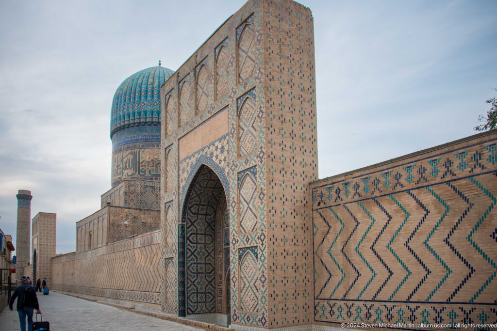 Angled view of a pointed side gate and long chevron-tiled wall with a turquoise dome beyond.