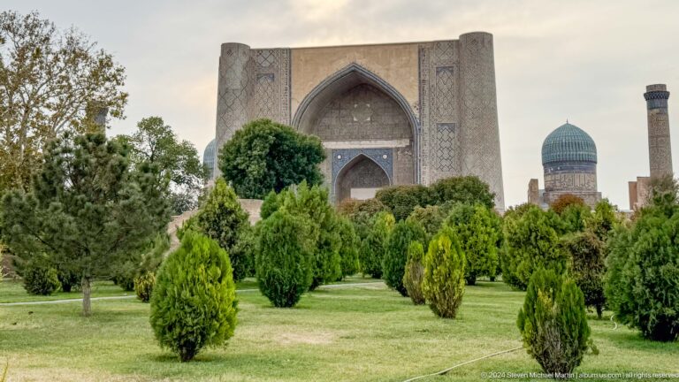 Symmetrical garden view of the monumental main iwan with shaped trees, lawn, and a turquoise dome at right.