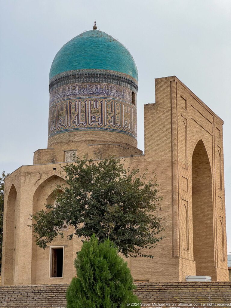 Decorated cylindrical tower beside a tall plain brick portal, partly obscured by a leafy tree.