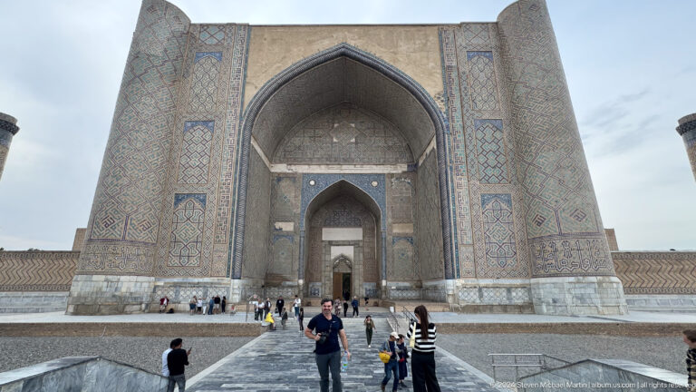 Frontal view of the enormous mosque iwan across a paved courtyard with scattered visitors.
