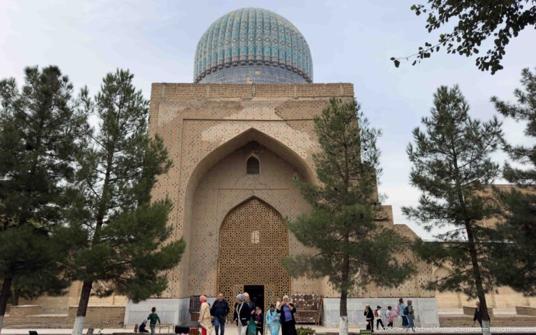 Domed brick mosque structure with a deep pointed portal, wooden lattice, trees, and visitors at the entrance.