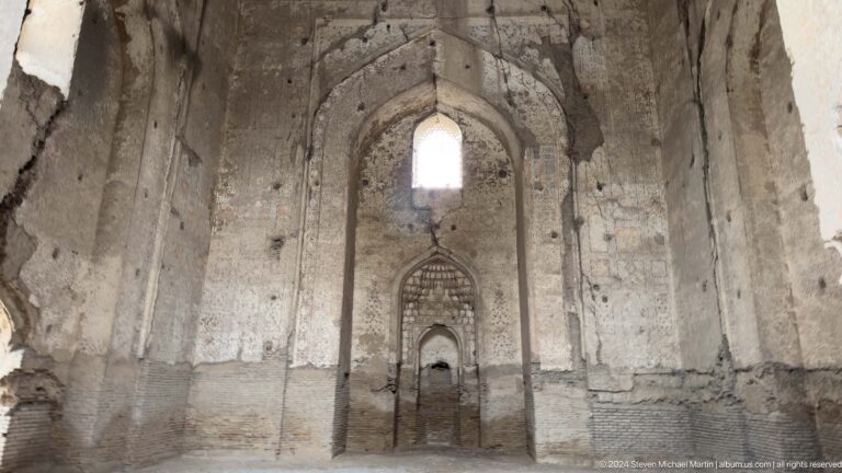 Broad ruined interior wall with a high pointed window, central niche, exposed brick, and worn plaster.