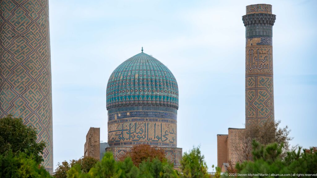 Turquoise mosque dome and tall decorated minaret rising above trees and low brick walls.