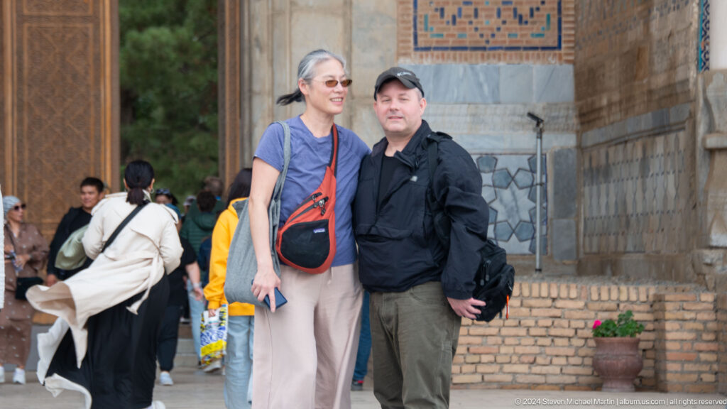 Two adult visitors smiling together beside a brick wall with blue tile decoration at the mosque complex.