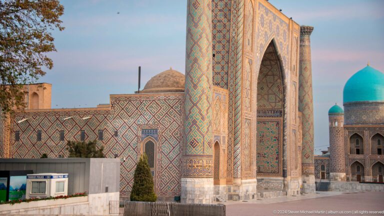 Side-angle view of Ulugh Beg Madrasa with a tall tiled portal and adjoining brick façade.