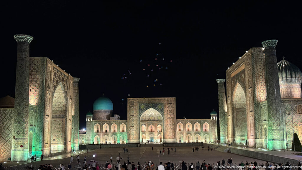 Night view of illuminated Registan Square with crowds below and small glowing lights rising into the sky.