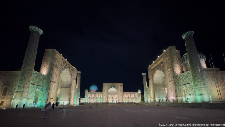 Low wide-angle nighttime view across nearly empty Registan Square toward illuminated Tilya-Kori Madrasa.