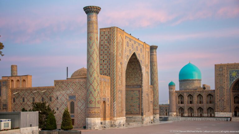Wide side view of Ulugh Beg Madrasa with portal, minaret, brick walls, and a turquoise dome beyond.