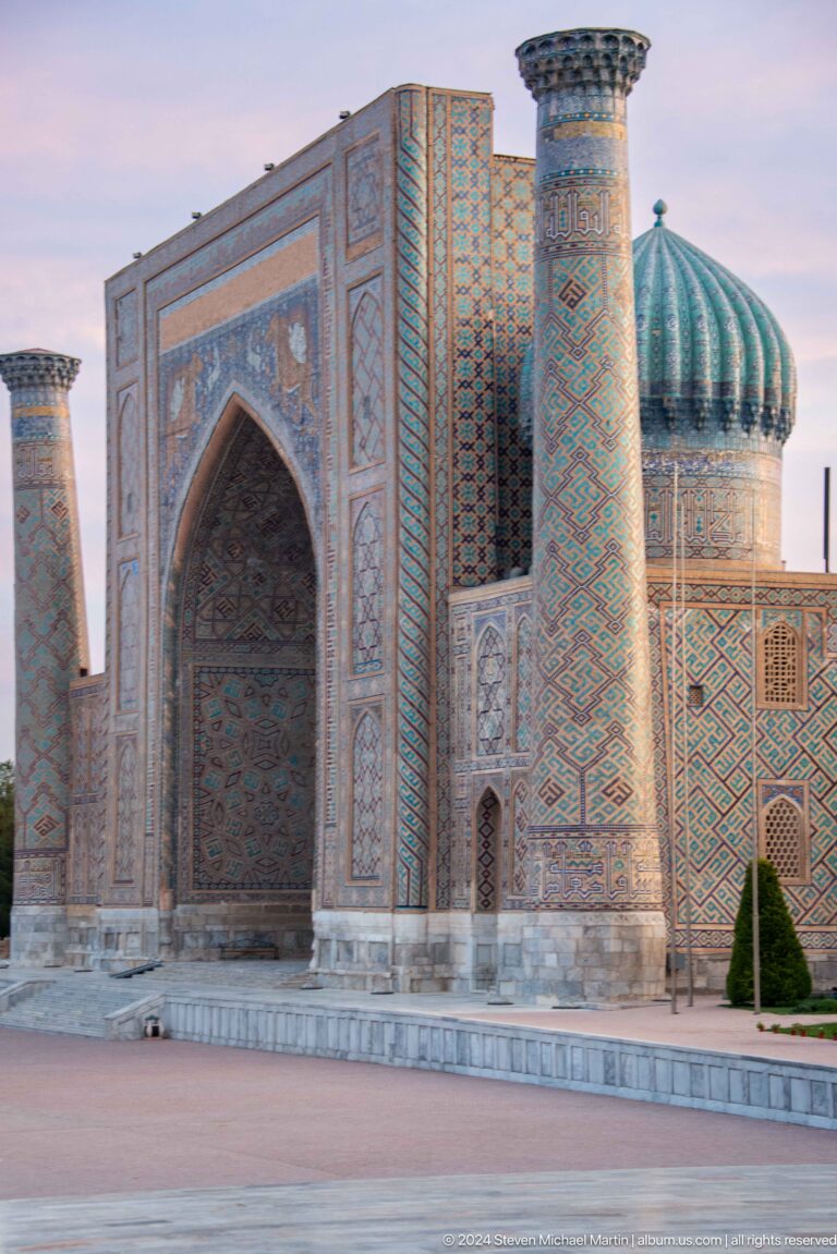 Shadowed pointed portal beside a tiled minaret and ribbed turquoise dome at Sher-Dor Madrasa.