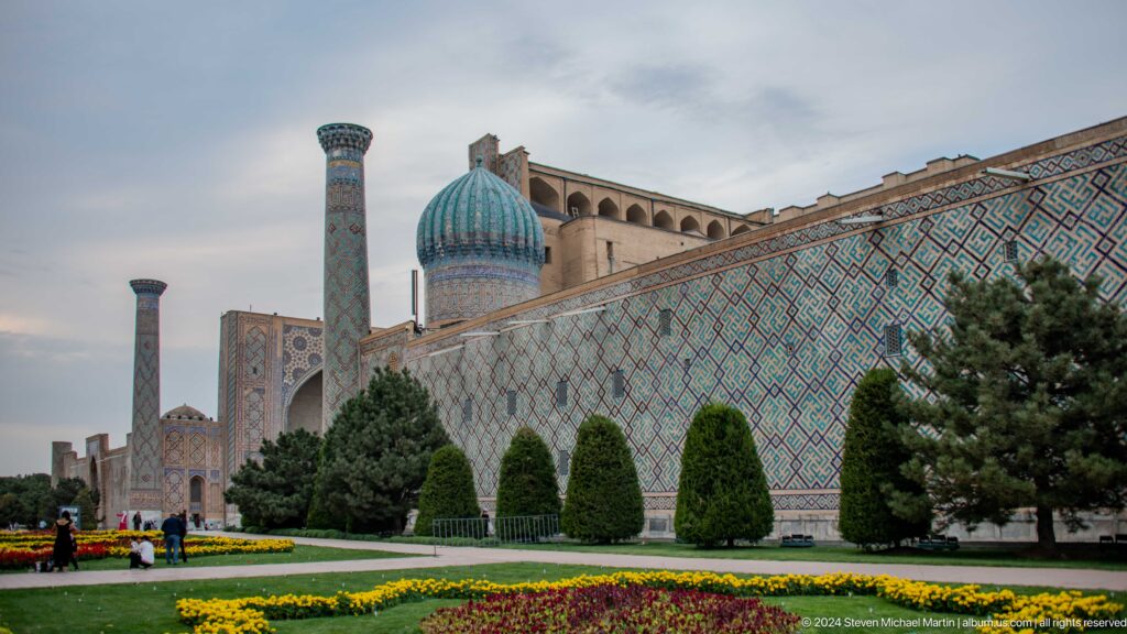 Angled view of a tiled Registan madrasa behind clipped shrubs, flowers, minarets, and a turquoise dome.