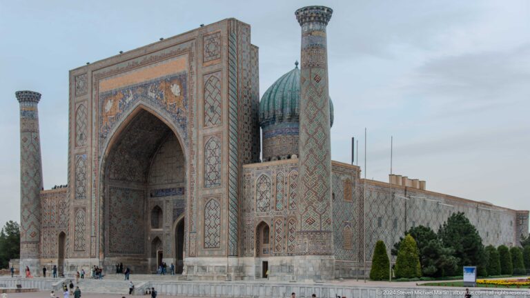 Angled view of Sher-Dor Madrasa with a monumental portal, minaret, and ribbed turquoise dome.