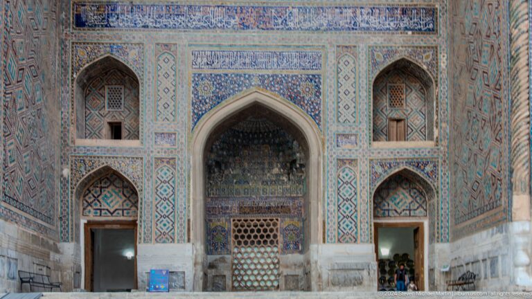 Central pointed doorway flanked by tiled arched niches on a Registan madrasa façade.