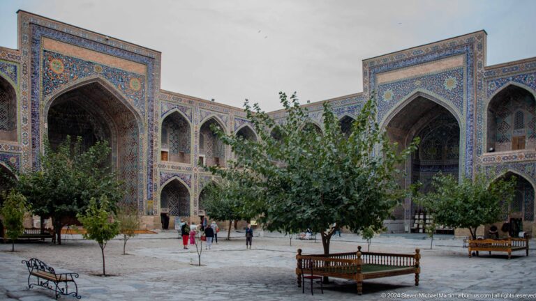 Tree-filled courtyard enclosed by tiled arcades with benches and visitors.