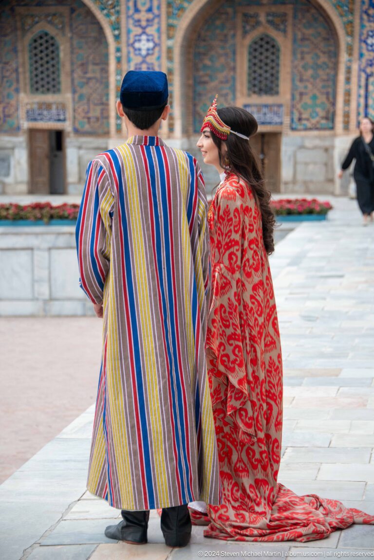 Two performers in long striped and red patterned robes standing in Registan Square.