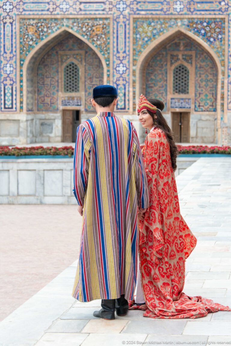 Two costumed performers in striped and red patterned robes speaking in Registan Square.