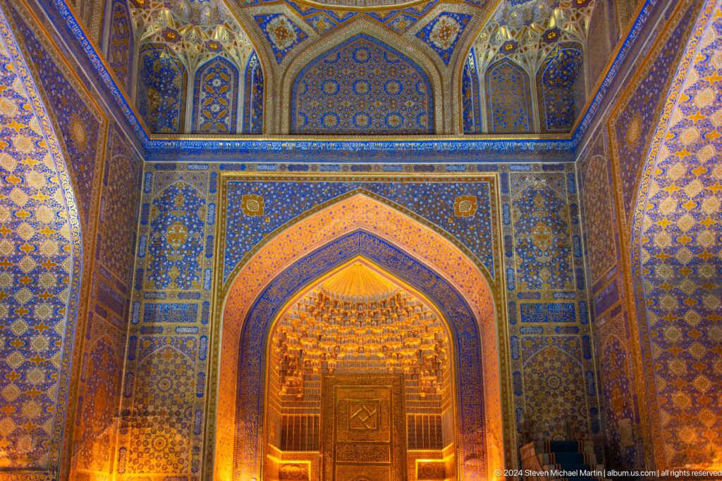 Blue-tiled interior with a deep pointed arch and warmly illuminated doorway at the Registan in Samarkand.