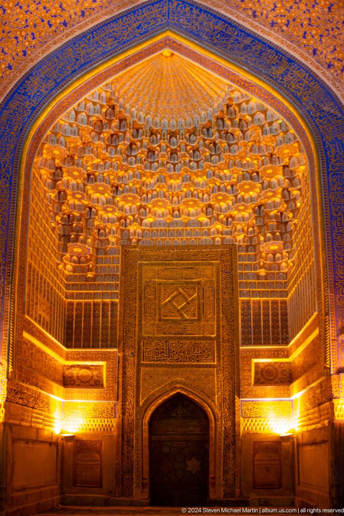 Golden-lit muqarnas canopy above a carved wooden double door inside the Registan complex.