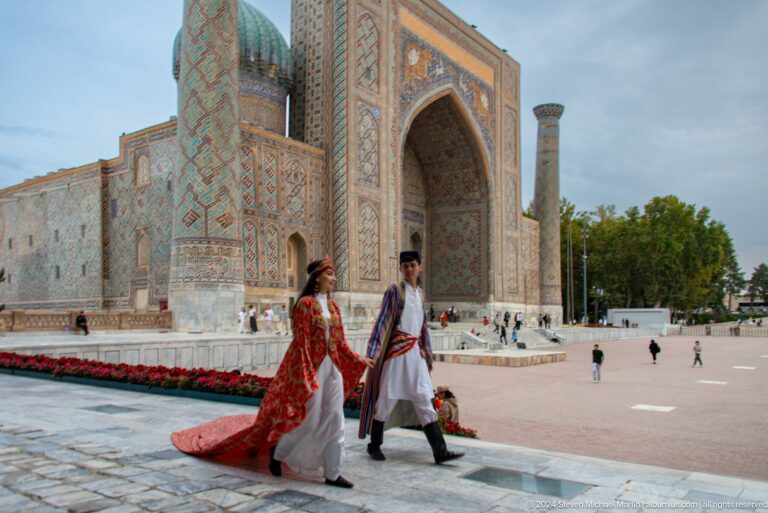 Two people in colorful traditional dress walk across Registan Square before a tiled madrasa.