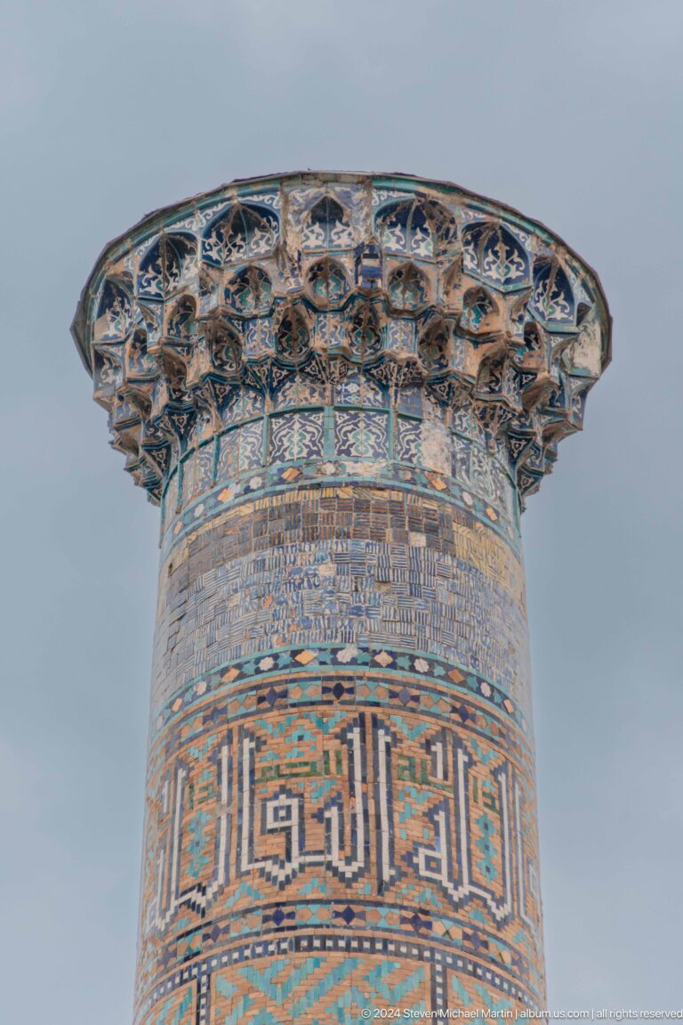 Upper section of a decorated Registan minaret with patterned brickwork, calligraphic bands, and a projecting crown.