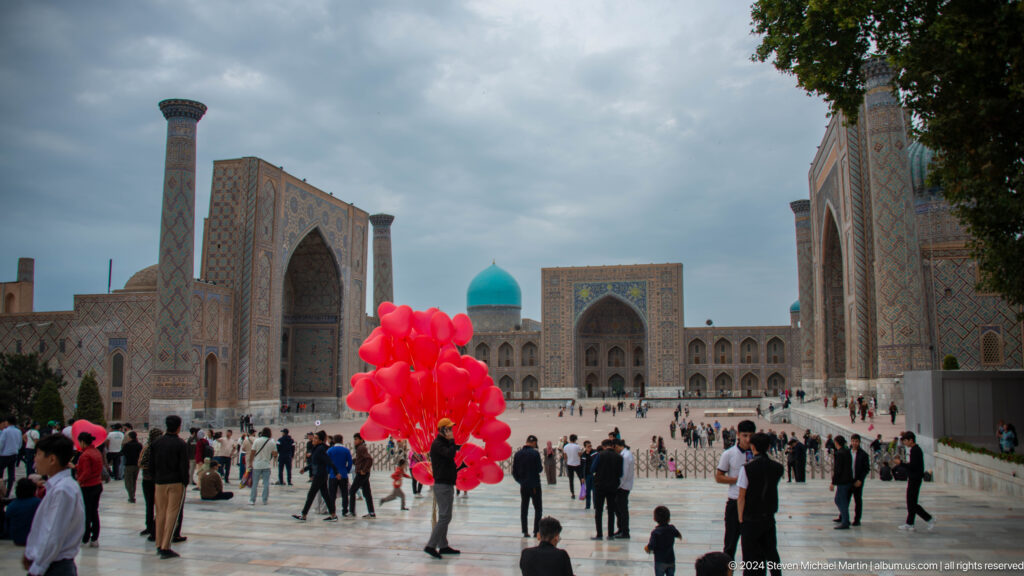 Vendor carrying bright red balloons through crowded Registan Square beneath the surrounding madrasas.
