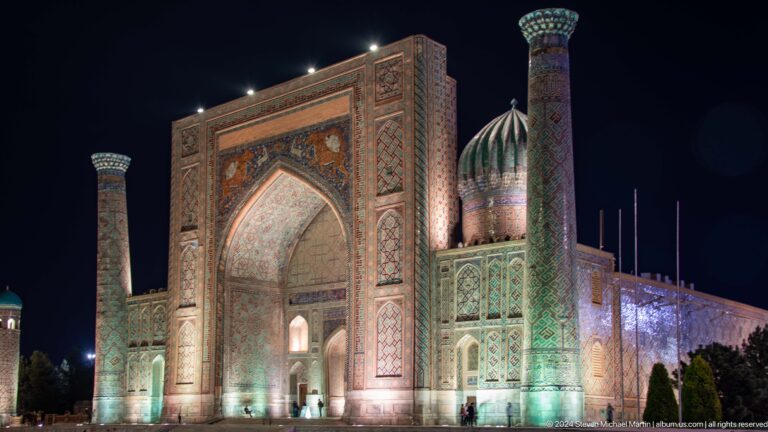 Oblique nighttime view of Sher-Dor Madrasa with monumental portal, ribbed dome, and minaret.