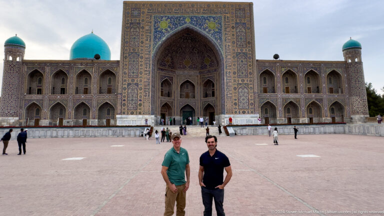 Two men posing in Registan Square with Tilya-Kori Madrasa and its turquoise dome behind them.