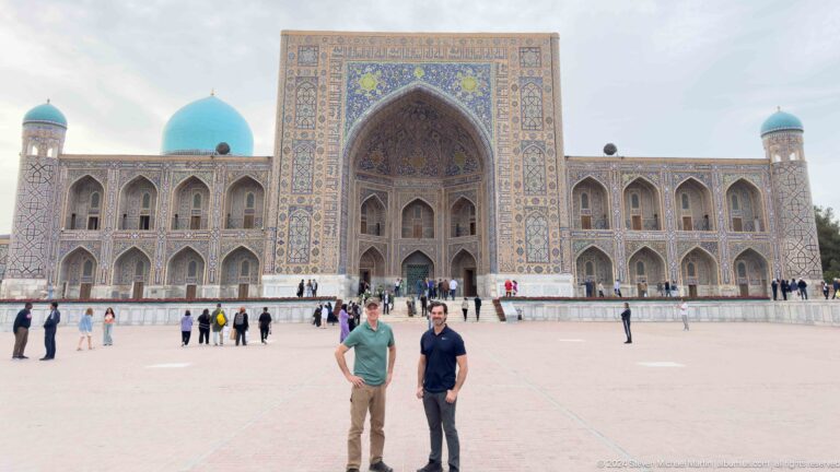 Two men standing in Registan Square before the full facade of Tilya-Kori Madrasa.