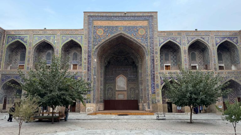 Courtyard facade with a tiled central iwan, two-story arches, mature trees, and paved ground.