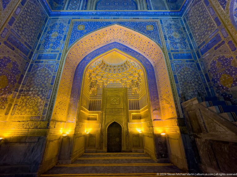 Blue-and-gold prayer hall interior with a tall pointed niche, calligraphy, and warm lighting.