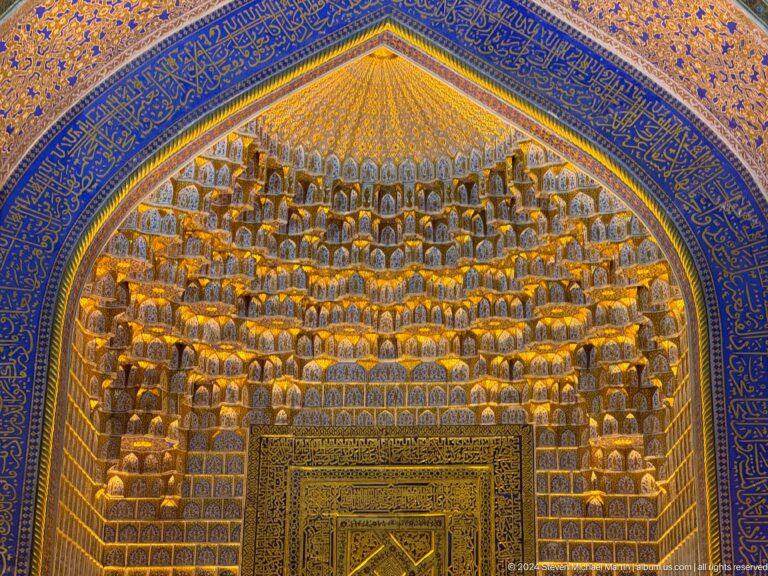 Golden tiered muqarnas filling a pointed vault above a framed doorway and blue inscription bands.