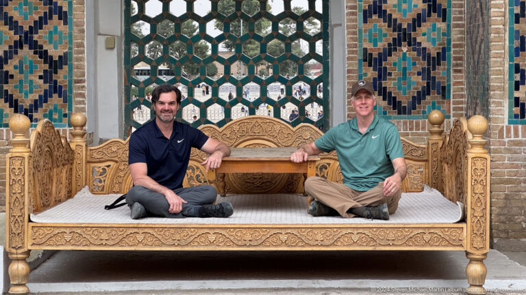 Two men seated cross-legged on an ornate carved wooden platform before a green lattice and tiled wall.